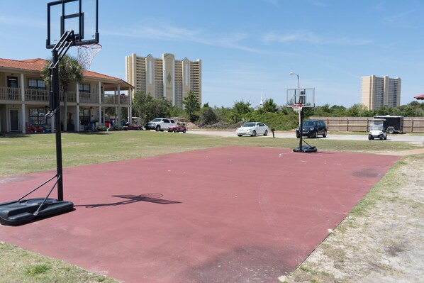An outdoor basketball court with a red surface, surrounded by parked cars and buildings in the background.