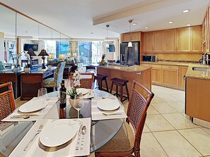 A modern kitchen with wooden cabinets, a central island, and a dining area with a glass table set for four. - Fully Upgraded Kihei Condo Sweeping Ocean Views Private Lanai Pool Hot Tub (Kihei)