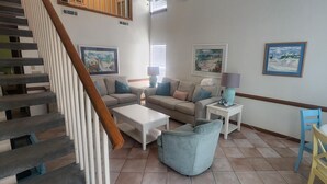 A spacious living area with a staircase, a sofa set, a coffee table, a side table with a lamp, and a dining table with chairs.