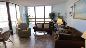 Una sala de estar con un sofá marrón, sillones, una mesa de centro de vidrio y vistas a un edificio a través de amplios ventanales.