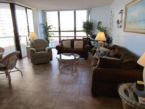 Sala de estar com sofá marrom, poltronas, mesa de centro de vidro e vista de um edifício através de grandes janelas.
