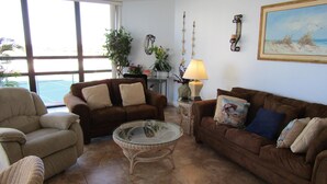Una sala de estar con un sofá marrón, un sillón beige y una mesa de centro de vidrio. Hay una lámpara sobre una mesita auxiliar y un cuadro en la pared.
