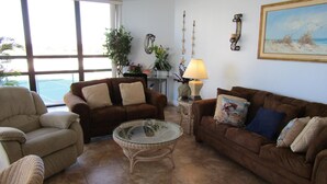 Una sala de estar con un sofá marrón, un sillón beige y una mesa de centro de vidrio. Hay una lámpara en una mesita auxiliar y un cuadro en la pared.