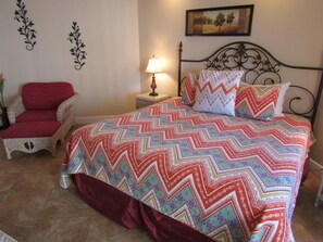Un dormitorio con una cama grande, una silla, una mesita de noche con lámpara y una pared con elementos decorativos.