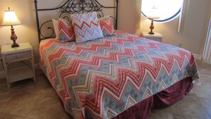 Una cama con una colcha de colores y un estampado en zigzag, dos lámparas de noche, un cuadro en la pared y una ventana con vistas.