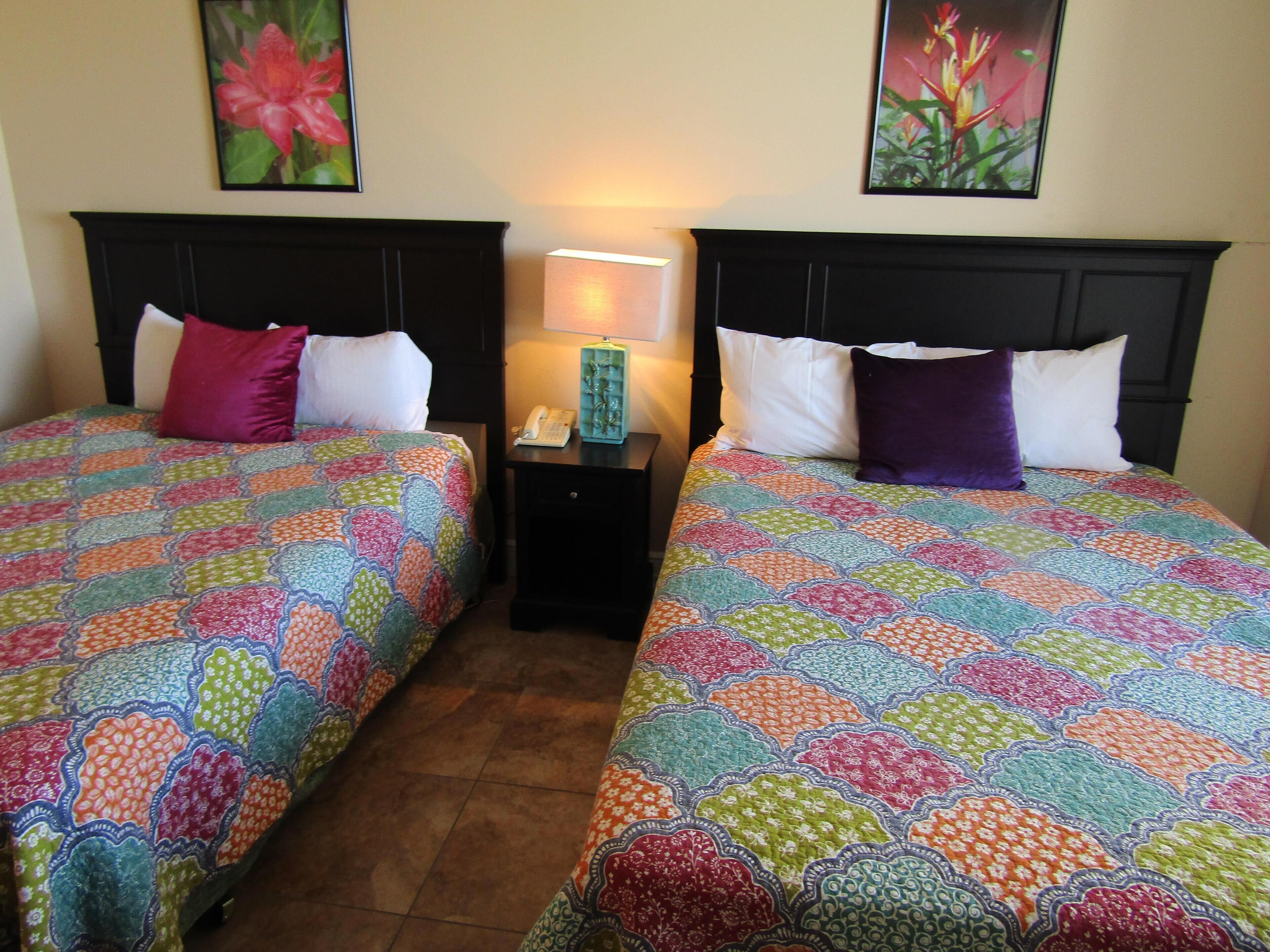 Deux lits doubles avec des couvertures aux motifs colorés, une table de chevet avec une lampe et des œuvres d’art florales accrochées aux murs.