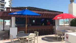 An outdoor bar area with tables and chairs, umbrellas, and a bar counter with a neon sign.