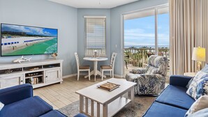 A living room with a TV, a dining table, chairs, a sofa, and a view of the beach. - Destin West Gulfside 410  ~ Amazing Beach View ~ Free Beach Service! (Fort Walton Beach)