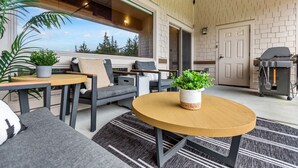 A modern outdoor seating area with a round wooden table, grey sofas, and potted plants.