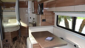 A compact RV interior with a bed, kitchenette, and a table with a tablet.