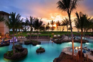 Un espace piscine aménagé dans un complexe hôtelier, avec des chaises longues, des palmiers et une vue sur le coucher de soleil.