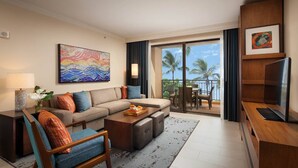 A modern hotel room with a sofa, armchair, and coffee table, a flat-screen TV, and a balcony with a view of palm trees.
