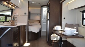 Interior compacto de una caravana, con cocina americana, cama y zona de comedor.