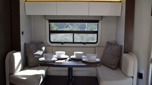 Interior compacto de una autocaravana con zona de comedor, dos asientos acolchados, una mesita y una ventana que ofrece vistas a la vegetación.