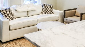 Una sala de estar moderna con un sofá blanco, un sillón estampado, una mesa de centro de mármol y un amplio ventanal con cortinas.