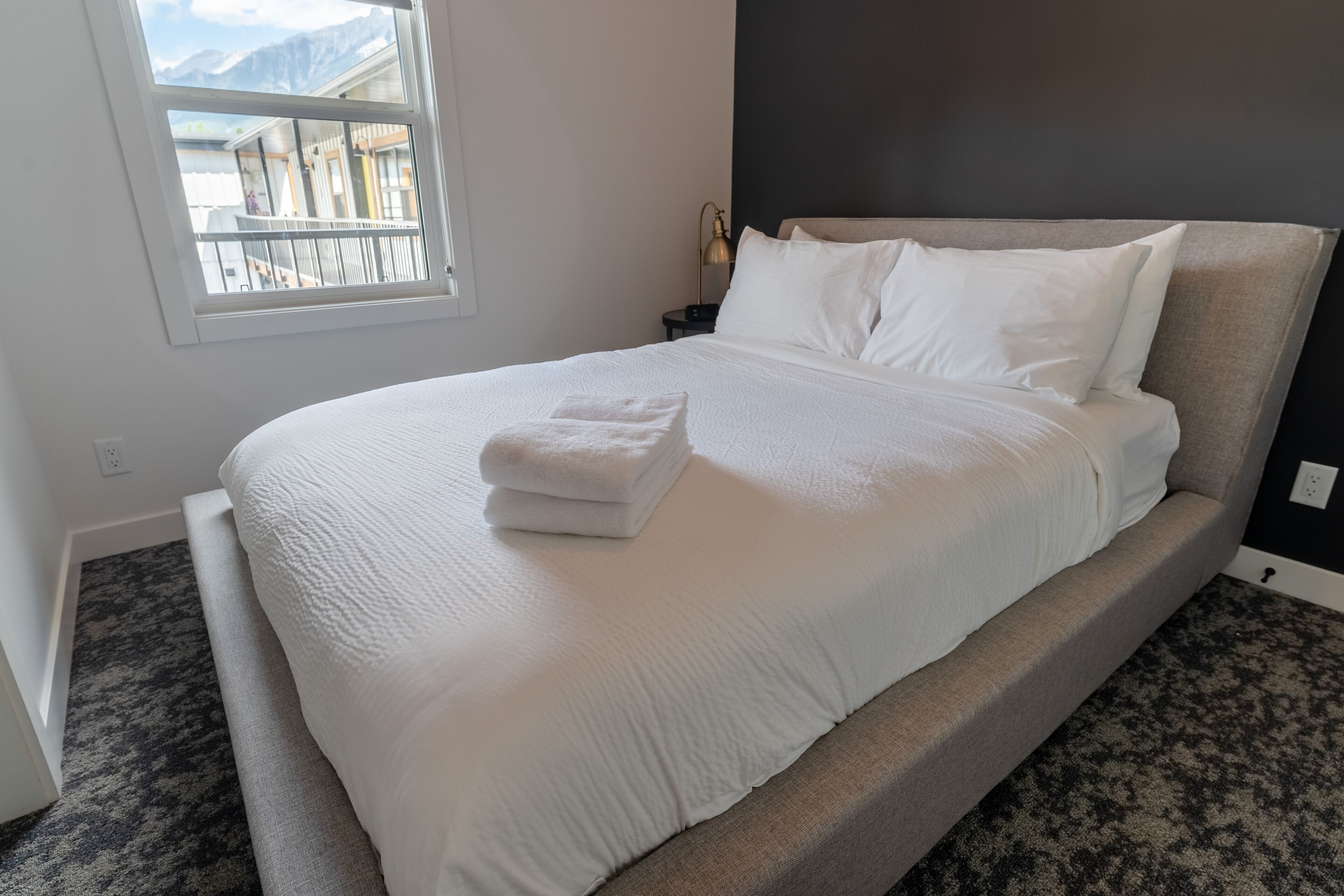 Una cama bien hecha con ropa de cama blanca y una toalla doblada, una lámpara de noche y una ventana con vistas a montañas.