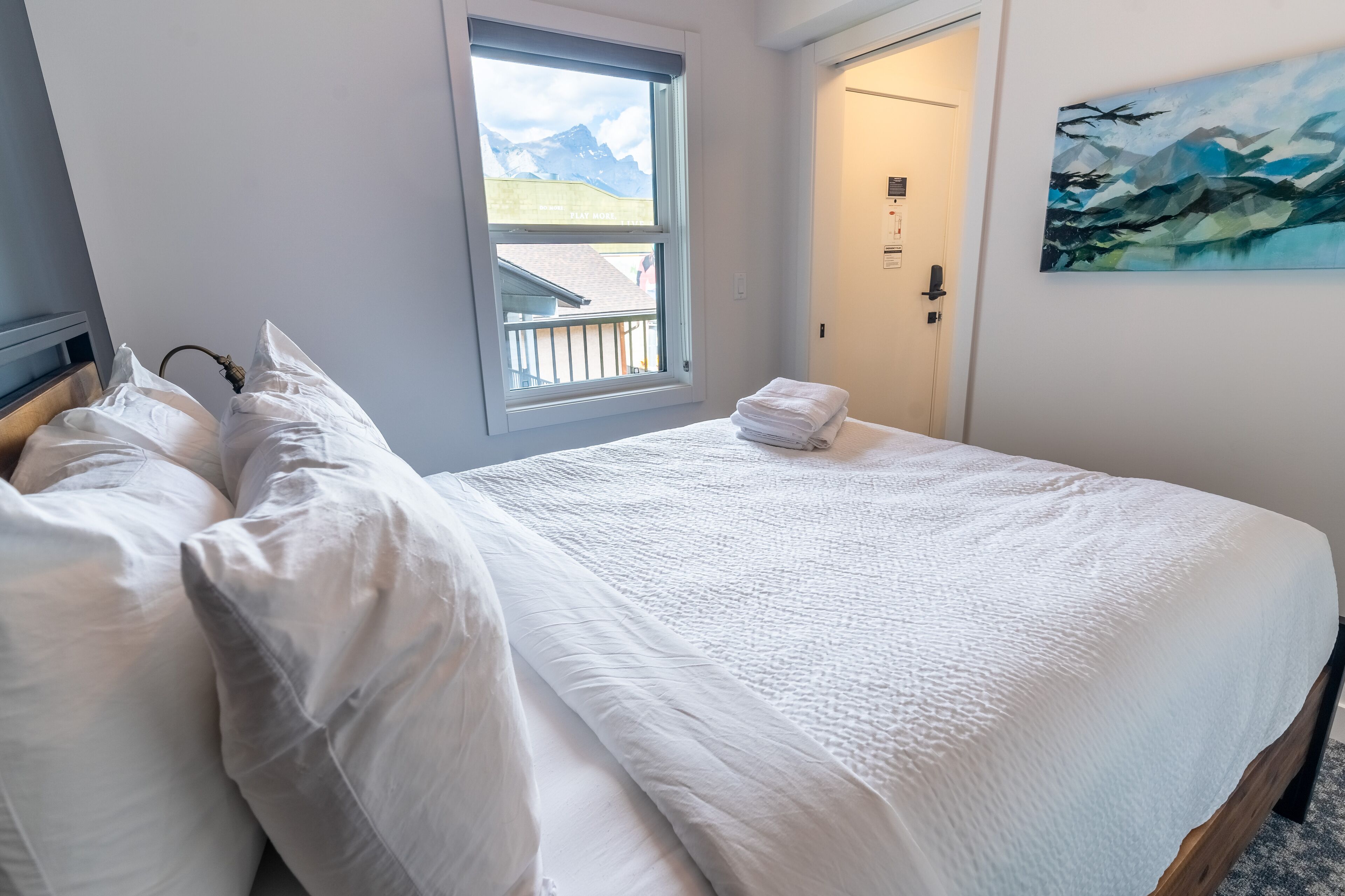 Een net opgemaakt bed met witte lakens en een gevouwen handdoek, een raam met uitzicht op bergen, en een schilderij aan de muur.