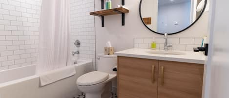 Une salle de bain moderne avec une baignoire blanche, des toilettes et un lavabo surmonté d’un miroir. Au-dessus du lavabo, on trouve des étagères en bois, et le sol est recouvert d’un carrelage à motifs noir et blanc.