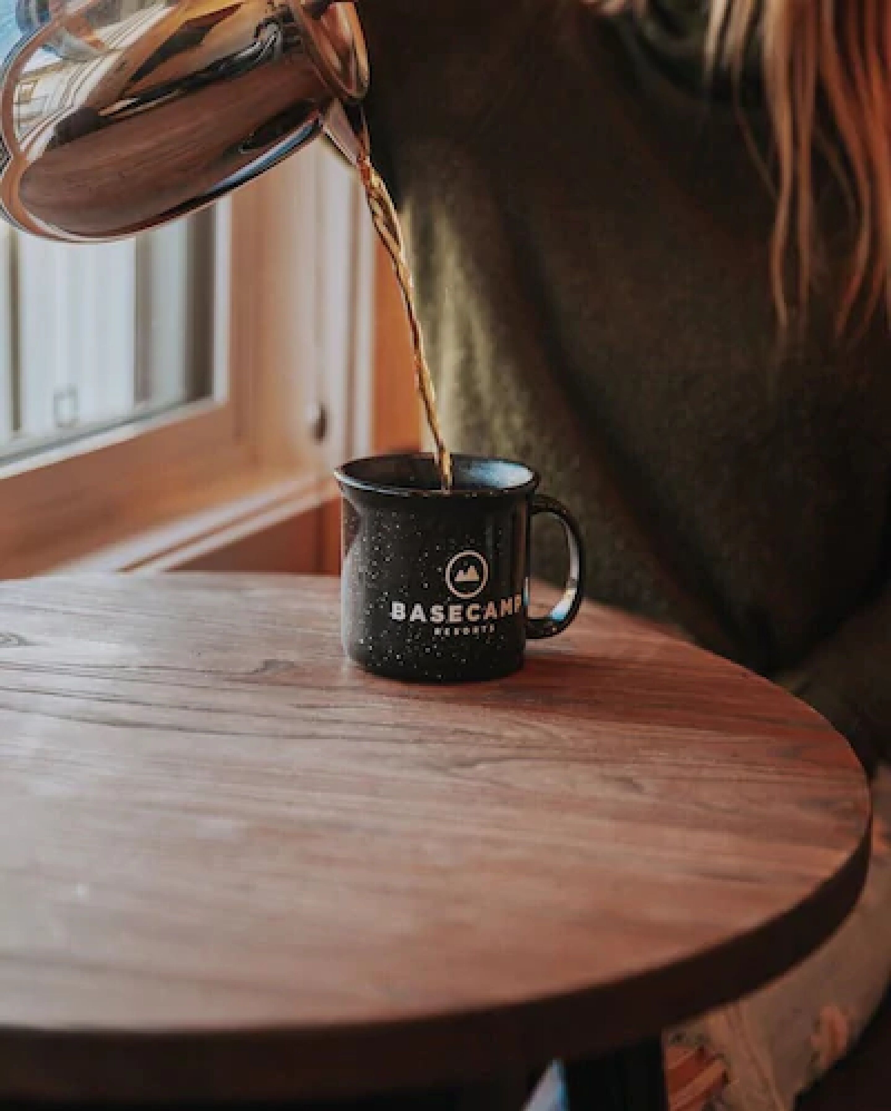 Una persona vertiendo un líquido desde un hervidor en una taza negra con el texto "BASECAN" grabado, sobre una mesa de madera.