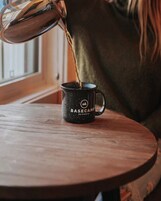 Una persona vertiendo un líquido desde un hervidor en una taza negra con el texto "BASECAN" grabado, sobre una mesa de madera.