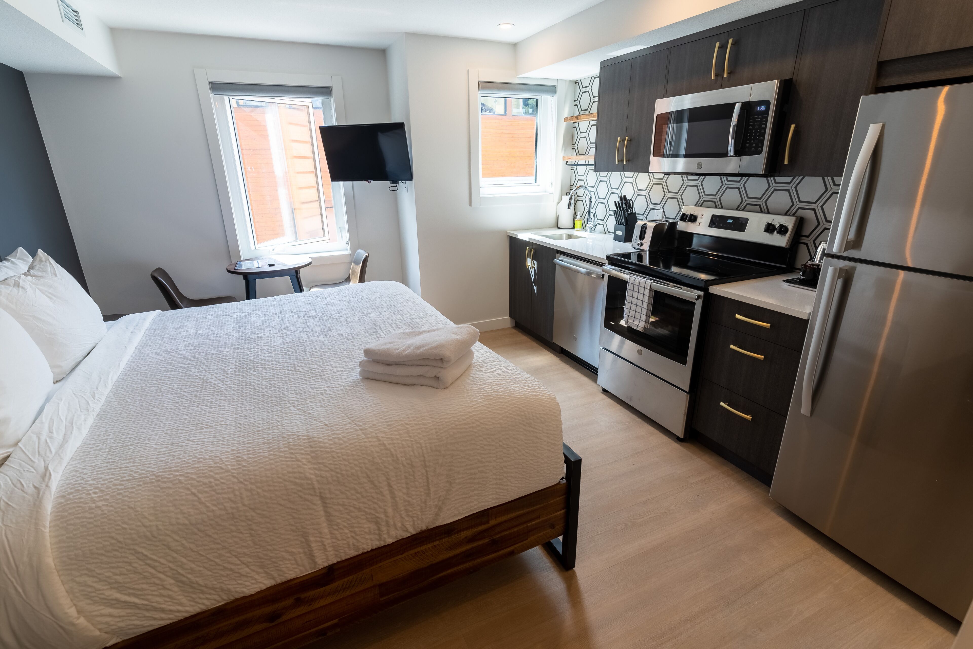 Habitación de hotel moderna con una cama grande, un escritorio pequeño, televisor de pantalla plana, cocina con electrodomésticos de acero inoxidable y ventana con vistas al exterior.