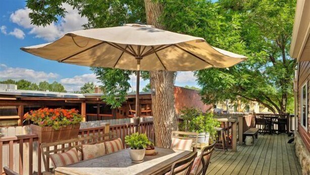 Een terras buiten met een houten tafel en stoelen, een grote parasol en potplanten.