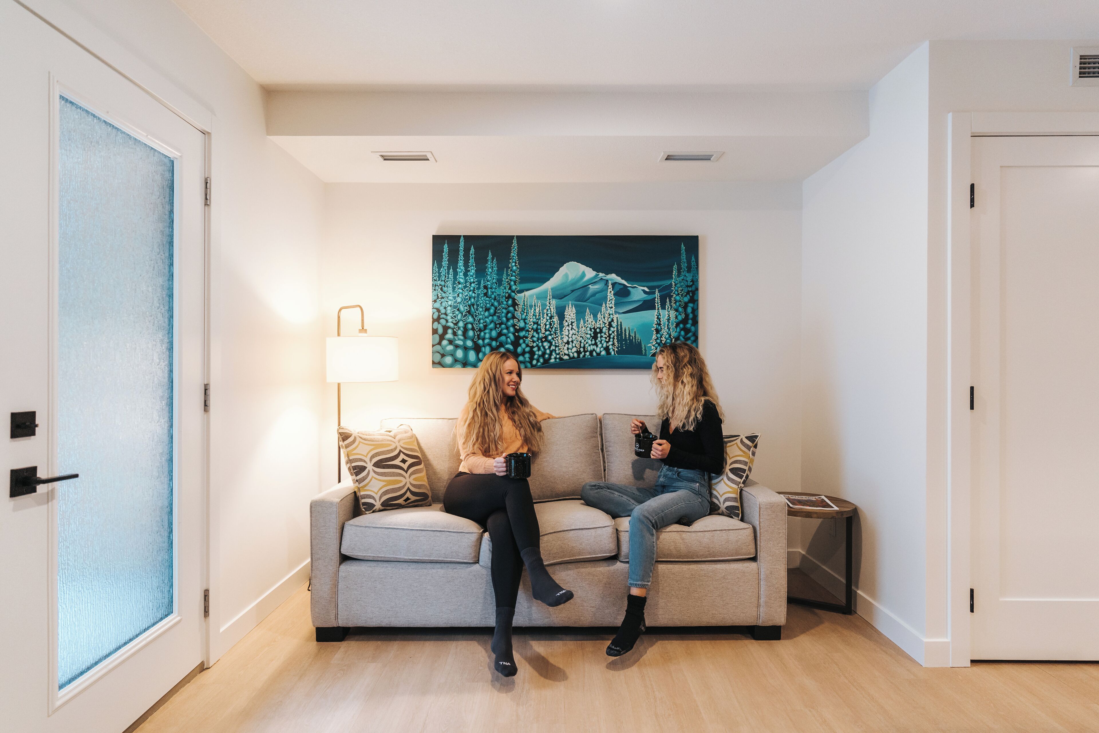 A room with a sofa, a painting of a mountain and forest, a lamp, and a door.