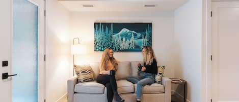 A room with a sofa, a painting of a mountain and forest, a lamp, and a door.
