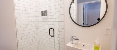 A modern bathroom with white subway tiles, a glass-enclosed shower, a round mirror, and a sink with a soap dispenser.