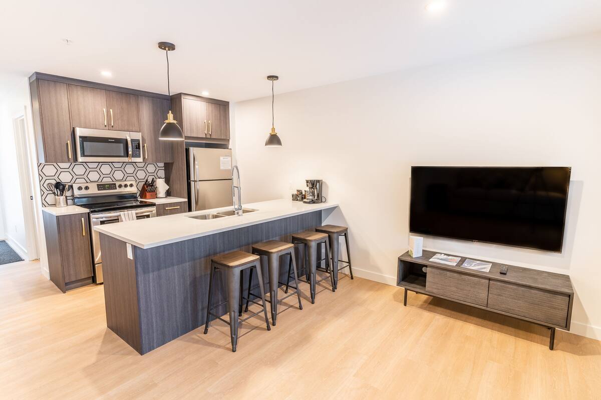 A modern kitchen with a central island, bar stools, a built-in microwave, and a large flat-screen TV mounted on the wall.