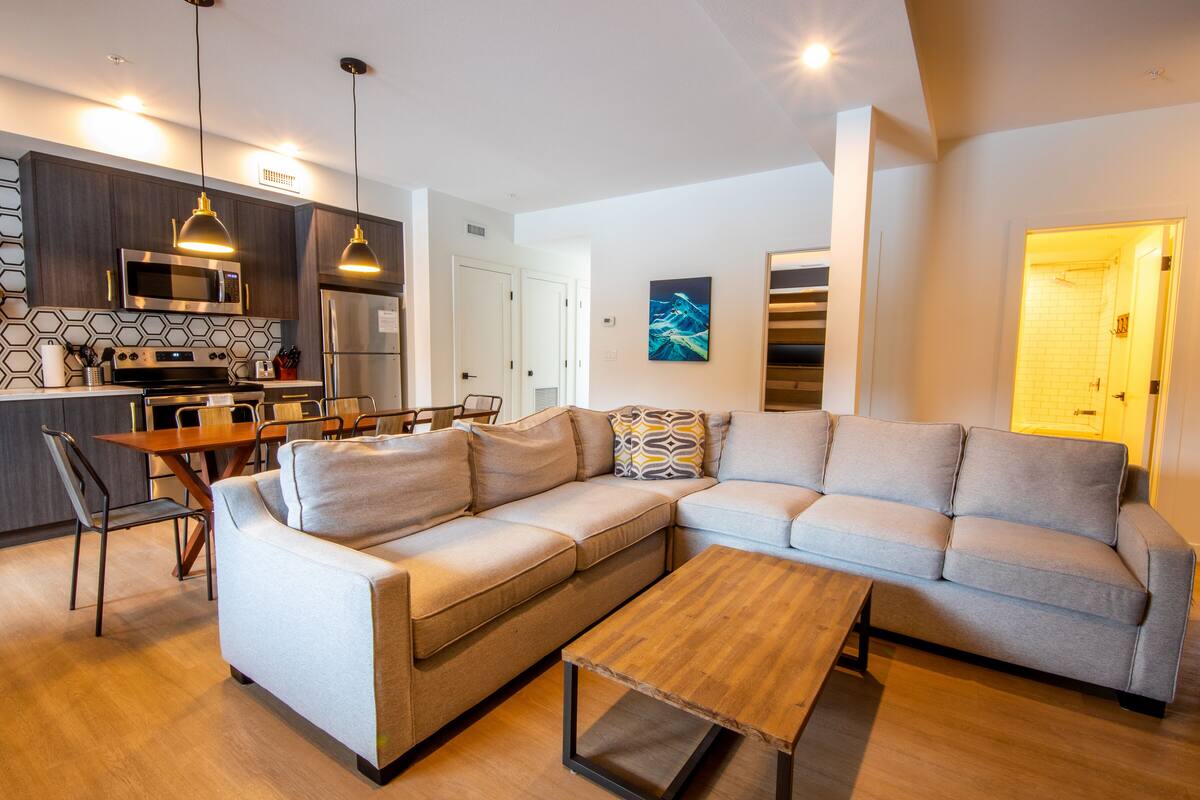 A modern living room with a sectional sofa, a wooden coffee table, and a dining area with a kitchen in the background.
