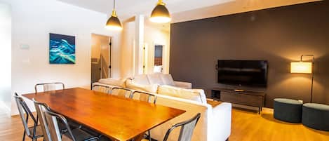 A modern living room with a wooden dining table, white sofa, and a flat-screen TV.