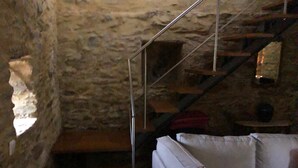 Ein Zimmer mit Steinwänden, einer Treppe zu einer oberen Ebene, einem grauen Sofa und einem kleinen Tisch mit einer Lampe.