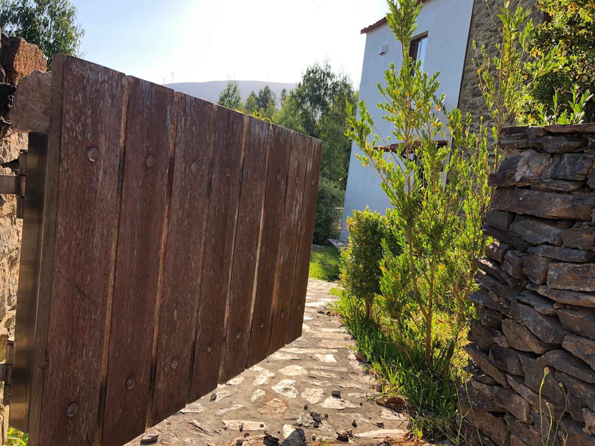A rustic wooden gate leading to a stone pathway, flanked by greenery and a white building with a stone wall.