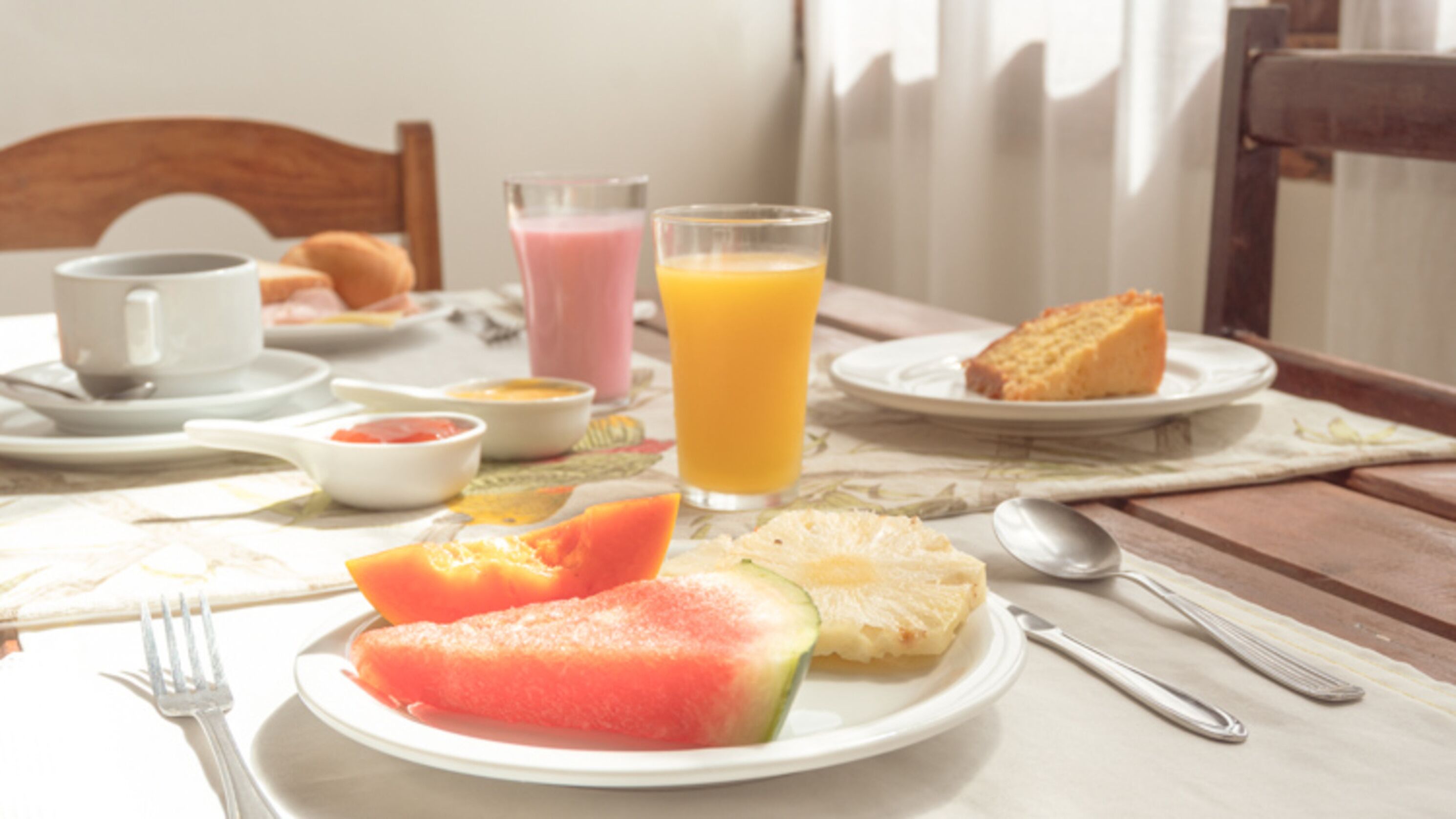 A breakfast table with plates of fruit, a glass of orange juice, and a slice of cake.