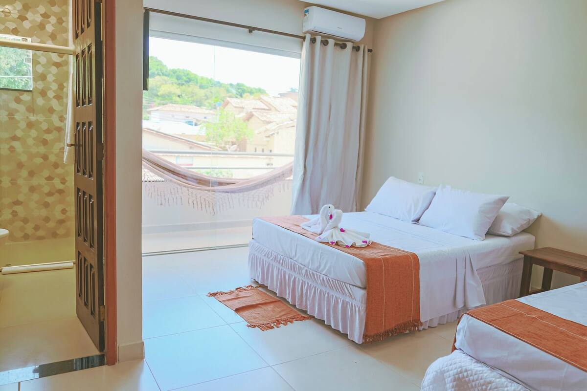 A hotel room with a bed, a small table, a window with a view of buildings, and a door.