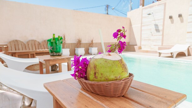 A wooden table with a basket containing a coconut and pink flowers, a green bottle, and a white bottle, with a pool and lounge chairs in the background. - Pousada Porto Da Barra (Porto Seguro)
