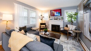 A living room with a sofa, armchairs, a coffee table, a fireplace, and a TV mounted on the wall. - Market 6min, HPU 2Min, Private Patio, Downtown 5min, Dogs Ok, 3 SMART TVs (High Point)