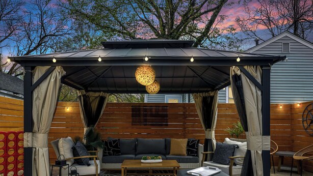 A covered outdoor seating area with a sofa, ottoman, and chairs, surrounded by wooden panels and a patterned rug. - Market 6min, HPU 2Min, Private Patio, Downtown 5min, Dogs Ok, 3 SMART TVs (High Point)