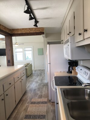 Una cocina con refrigerador, estufa y microondas. Hay una ventana con persianas y una puerta que da a otra habitación.