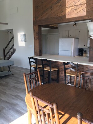 Un comedor con mesa y sillas de madera, una cocina al fondo y una escalera que lleva a otro nivel.