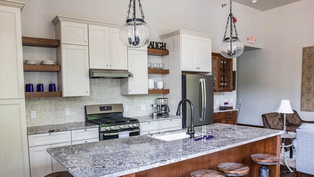 Una cocina moderna con armarios blancos, encimera de granito, refrigerador de acero inoxidable y estufa a gas. Hay taburetes de madera en la isla y una lámpara colgante arriba.