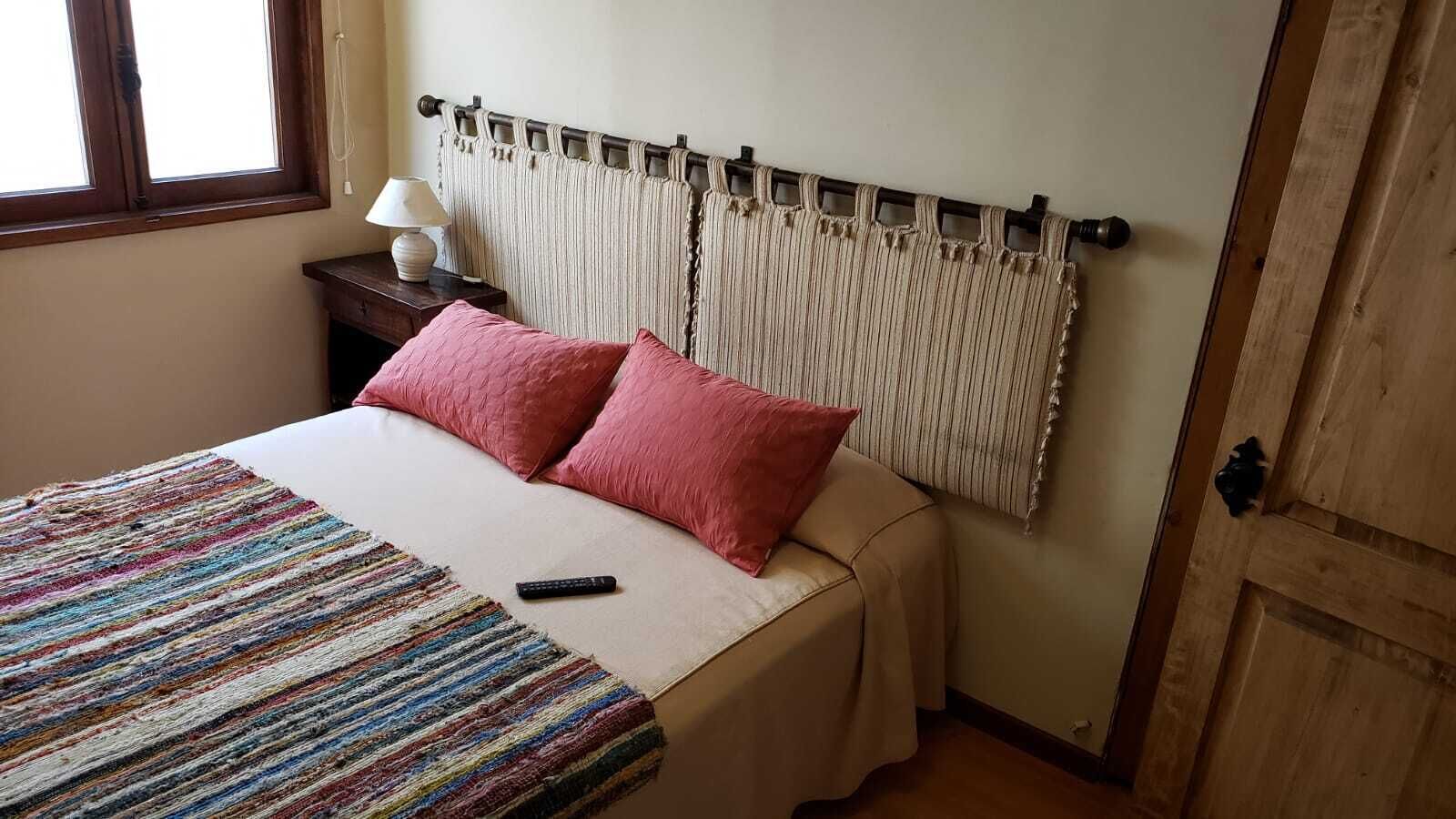 Una cama con un edredón a rayas, dos almohadas rosas, un control remoto y una mesita de noche con una lámpara.
