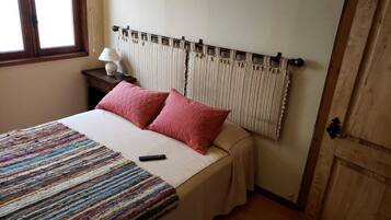 Una cama con un edredón a rayas, dos almohadas rosas, un control remoto y una mesita de noche con una lámpara.