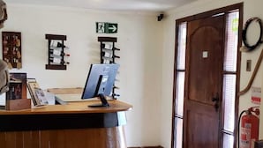 A reception area with a wooden reception desk, a computer, and a fire extinguisher. There is a door with a window and a wall-mounted emergency exit sign. - Tierra y Vino (Santa Cruz)