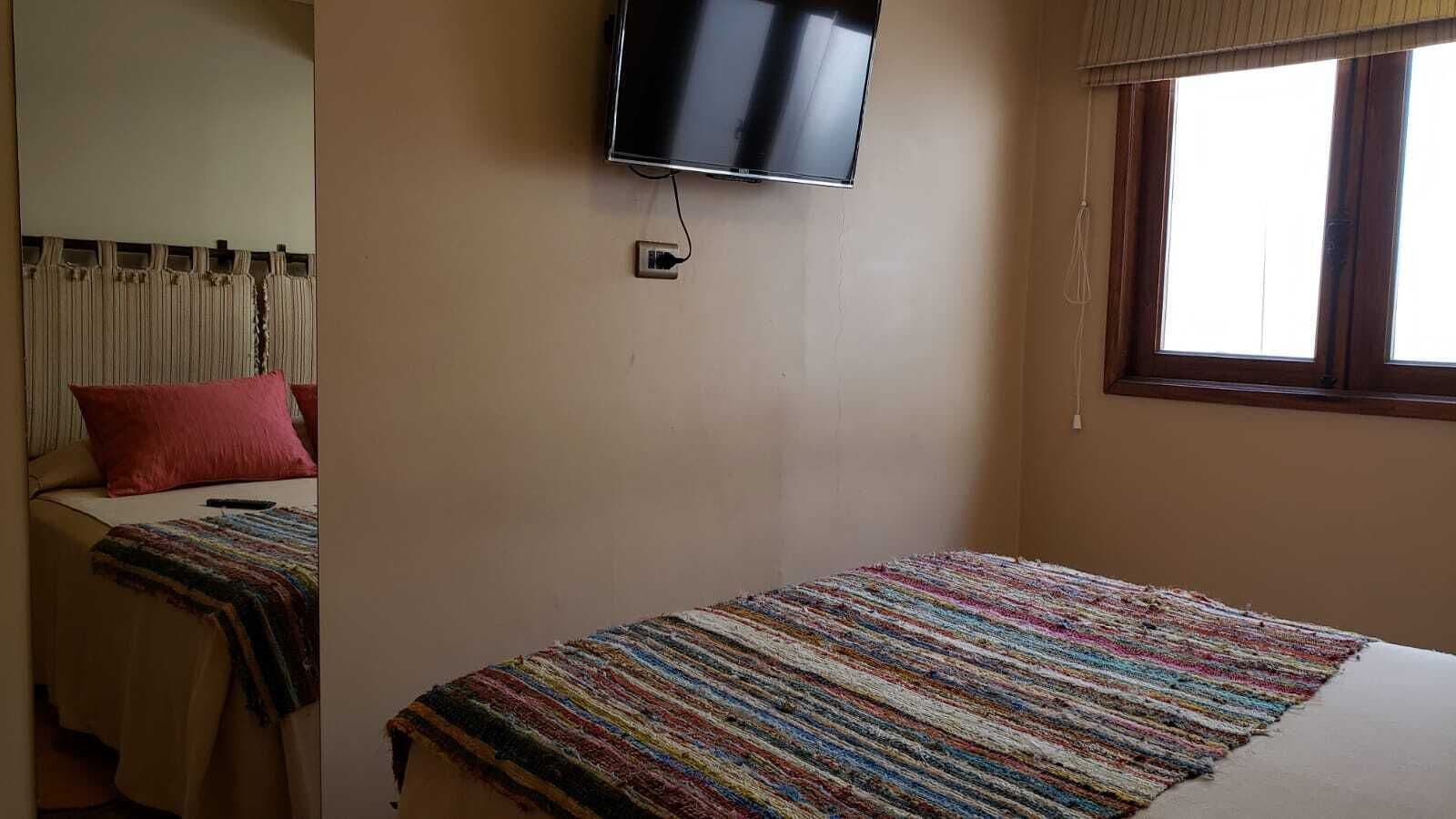A single bed with a colorful striped blanket, a television mounted on the wall, and a window with curtains.