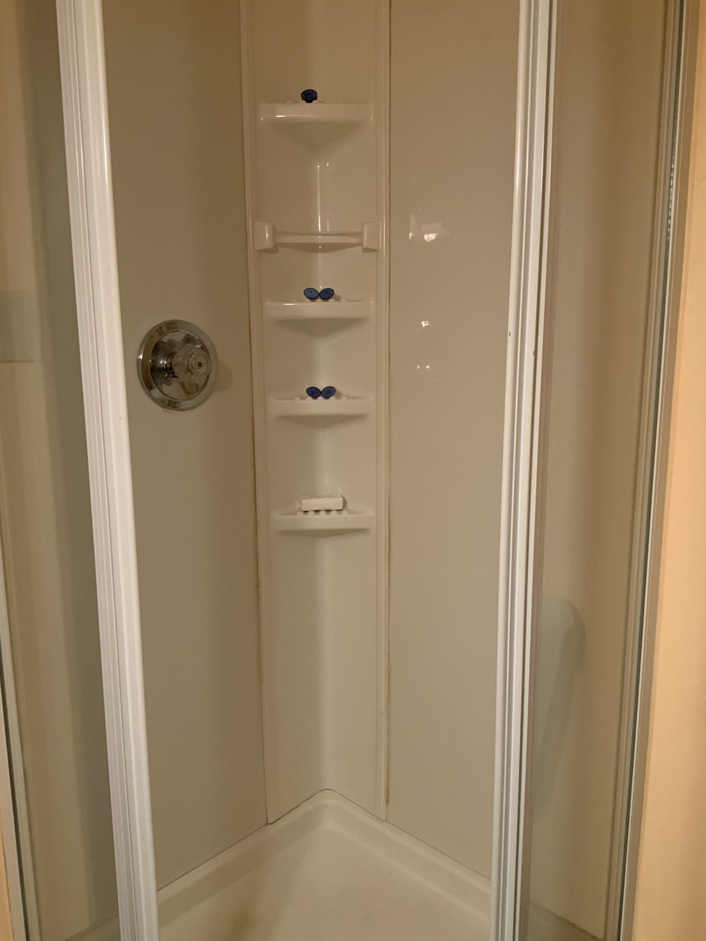 Une douche d’angle avec une porte en verre, un mur blanc et un sol blanc.