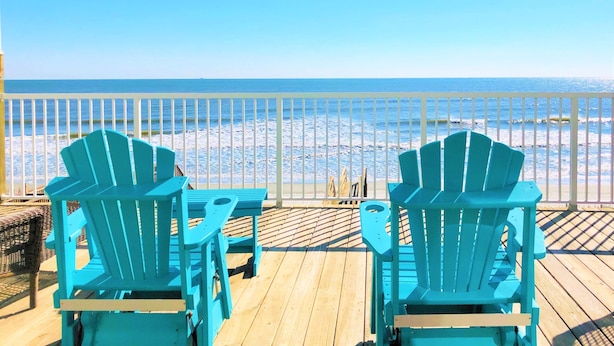 Une terrasse en bois avec des fauteuils Adirondack turquoise, donnant sur une plage.