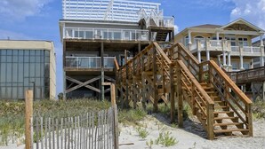Une propriété située en bord de mer, dotée d’un escalier en bois menant à un balcon, d’une zone clôturée et d’un bâtiment avec de grandes fenêtres.