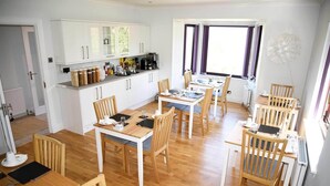 A modern kitchen with white cabinets, a dining area with wooden chairs and tables, and a large window with curtains. - Glendaruel Bed & Breakfast (Portree)